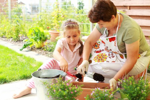 A woman and a young girl are working together on a garden planting project outdoors in a backyard. The woman is wearing a floral apron and gardening gloves, leaning over a rectangular plant bed filled with dark soil, while the girl, dressed in a light pink top and striped leggings, is holding a small garden trowel and assisting in planting. Surrounding them are vibrant green plants and potted flowers, with a well-maintained lawn and a wooden garden fence in the background. The garden scene appears to be during daylight, with natural light illuminating the area, and the environment suggests a neat, organized outdoor space ideal for gardening activities and lawn care services, possibly in Barking or nearby areas in London. This outdoor setup demonstrates typical gardening work involving planting and soil preparation, aligning with professional landscaping and garden maintenance offerings. The image emphasizes family-friendly gardening efforts in a healthy, lush garden environment, supported by a local landscaper such as Landscapers Barking.