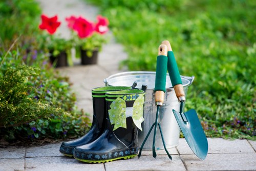 The image displays a tidy outdoor garden scene in Barking, showing a paved pathway with a pair of well-maintained black rubber gardening boots with green and beige patterned liners placed on the ground. Near the boots, there are a small metal bucket, a trowel, and a hand fork, all positioned for gardening tasks. In the background, a blurred garden with vibrant pink flowers in pots and lush green foliage is visible, indicating a cultivated and cared-for outdoor space. The scene suggests a moment of gardening activity or preparation, with natural light illuminating the setting, typical of a mild, fair-weather day suitable for outdoor gardening work. This arrangement and the visible tools reflect a service focus on garden maintenance, landscaping, and lawn care, echoing the expertise and offerings of Landscapers Barking in local residential gardens, possibly within the postcode area associated with Barking. The overall composition emphasizes a well-kept garden environment designed for planting, pruning, and outdoor aesthetic enhancement, aligning seamlessly with gardening services provided in the local area.