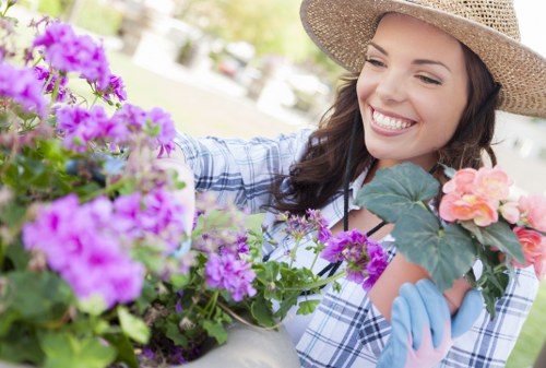 The image shows a smiling woman wearing a wide-brimmed straw hat and gardening gloves, tending to vibrant flowering plants in a well-maintained garden. In the foreground, there are lush purple and pink blossoms, with the woman appearing to be carefully inspecting or arranging the plants. The garden features a neatly trimmed lawn with dense greenery, including a backdrop of bushes and trees that provide natural shade. The scene is brightly lit with natural daylight, suggesting a sunny day with clear weather. The garden environment appears organized, with flower beds and a paved area visible, indicating attention to outdoor landscaping and planting. This setting exemplifies typical garden maintenance and planting practices that landscaping companies like Landscapers Barking may support, emphasizing a healthy, thriving outdoor space in a residential backyard near Barking or the East London area.