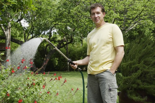 A man in a light yellow t-shirt and grey cargo pants is standing in a well-maintained garden, watering a flower bed with a garden hose attached to a spray nozzle. The garden features a lush, green lawn with a dense border of bushes and trees in the background, providing a natural and vibrant outdoor space. The flower bed contains small, bright red flowers growing amidst green foliage, bordered by a neat edge that separates it from the surrounding grass. Behind the man, there are mature trees with leafy branches and a textured hedge or shrubbery, contributing to a private and scenic environment typical of residential gardens in Barking. The scene is set under natural daylight, suggesting a clear, pleasant day suitable for outdoor garden maintenance. This type of gardening activity aligns with professional lawn and garden care services offered by Landscapers Barking to support healthy plant growth and attractive outdoor spaces in local homes.