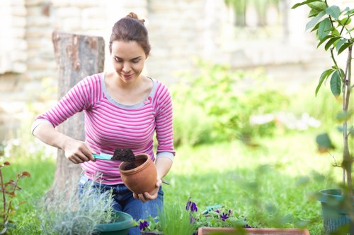 A young woman with dark hair tied back, wearing a pink and white striped long-sleeve top, is engaging in gardening activities in an outdoor back garden space. She is holding a terracotta flowerpot filled with dark soil in one hand while using a small green trowel with the other hand to transfer soil into the pot. The garden environment around her includes a neatly maintained green lawn in the foreground, and in the background, there are various plants and shrubs, some with purple flowers, alongside a weathered tree stump and a stone or brick wall that encloses the yard. The scene is illuminated by natural daylight, suggesting a mild, overcast day, creating a soft, natural light that highlights the vibrant greens of the grass and foliage. This outdoor space appears well-kept, suitable for gardening and landscaping projects, with a focus on planting and cultivating flowers or small plants, aligning with professional gardening and landscaping services offered by companies like Landscapers Barking. The setting reflects a typical residential backyard conducive to garden renovations and outdoor beautification in Barking, Essex.