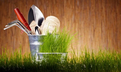 This image features a close-up view of a patch of lush, green grass in the foreground, with a wooden surface as the background. Centered in the image is a small, metallic gardening container filled with various garden tools, including a trowel, a hand rake, and a gardening fork, alongside a ball of twine and a clump of fresh, bright green grass blades. The tools are clean and well-organized, suggesting recent gardening activity. The wooden background provides a warm, natural tone that complements the vibrant green of the grass and the metallic sheen of the tools, evoking a healthy, well-maintained outdoor garden space typical of residential gardens in Barking. The overall scene highlights the preparatory or maintenance activities associated with gardening services, such as lawn care, planting, and general outdoor upkeep. The image demonstrates a professional and tidy gardening setting, ideal for a gardening or landscaping website aiming to showcase practical garden management in a suburban or urban outdoor environment.