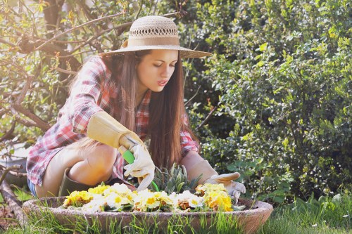 A young woman gardening outdoors in a backyard garden with lush green foliage and flowering plants, working at a low, circular flower bed bordered with soil and small rocks. She is wearing a wide-brimmed straw hat, a red and white checkered shirt, and gardening gloves while carefully planting or tending to yellow and white flowers. In the background, densely packed hedges and trees provide shade and natural privacy, with sunlight filtering through the leaves, creating a bright and inviting environment. The scene displays well-maintained grass and a variety of garden elements, characteristic of a landscaped outdoor space in Barking or a nearby area, highlighting professional gardening and lawn care services like those offered by landscapersbarking.co.uk.
