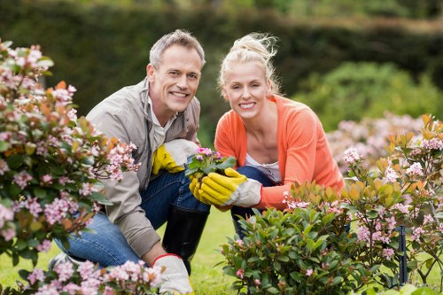 A man and a woman with light skin are gardening in a well-maintained front garden in Barking, with neatly trimmed flowering shrubs and lush green grass visible around them. The man, wearing a grey shirt, and the woman, dressed in an orange top, are both smiling at the camera while kneeling on the grass. They are holding a small potted plant between them, and the woman is wearing yellow gardening gloves. Behind them, the garden features a backdrop of trees and a hedge line, with a mixture of flowering plants and foliage adding colour and texture to the outdoor space. The scene is set during daytime with natural light, likely on a clear day, highlighting the vibrant colours of the flowers and foliage. This image relates to gardening services provided by Landscapers Barking, illustrating the care and attention involved in landscape and garden maintenance within local residential outdoor spaces, emphasizing lawn care, plant upkeep, and garden tidiness.